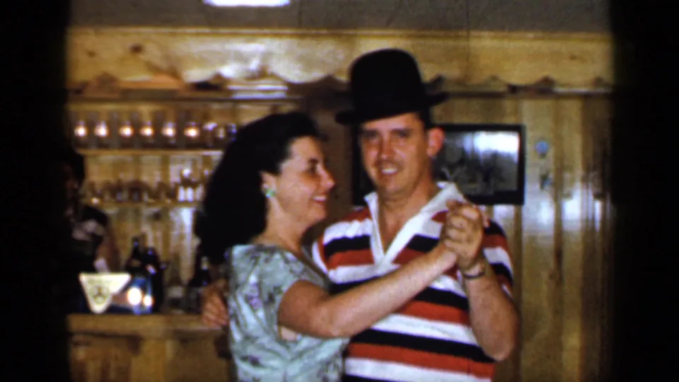 A man in a striped polo and dark hat dances with a smiling woman in a floral dress in the 1965 Long Island bar interior.