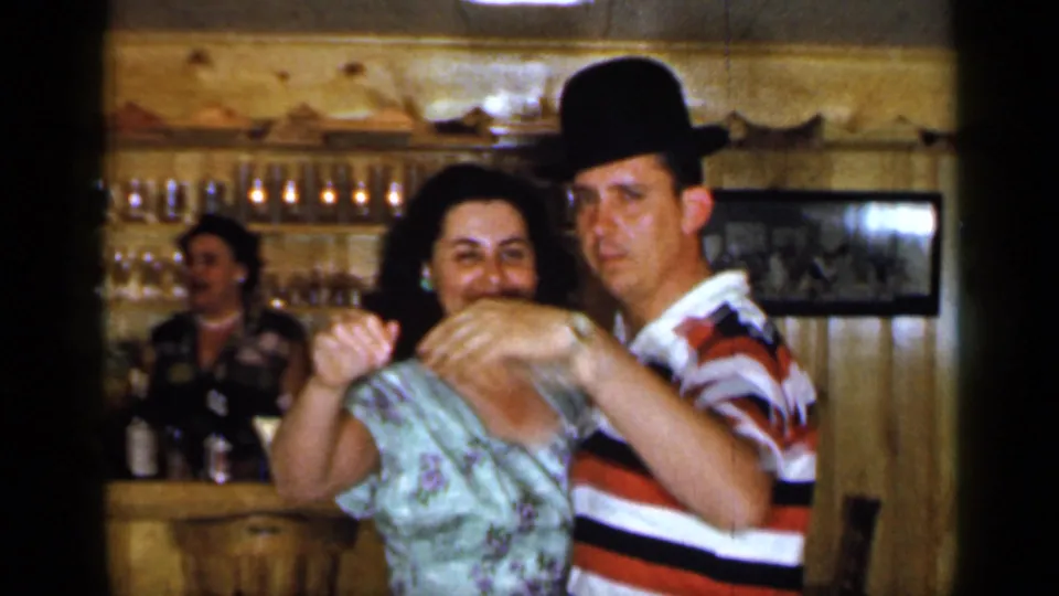 The dancing couple passes close to the camera as another adult stands behind the bar in this 1965 Long Island home-movie frame.