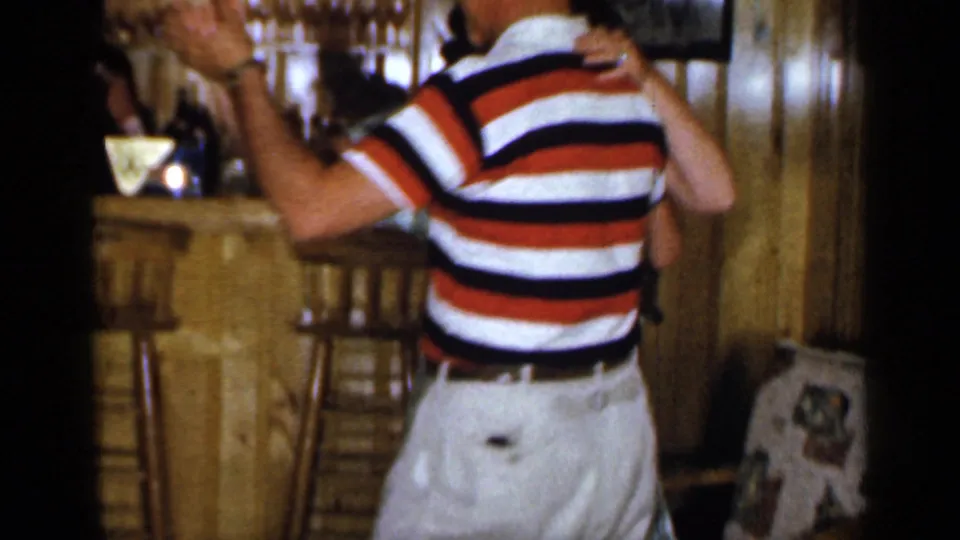 In 1965 Long Island home-movie footage, an adult couple dances closely near a wood-paneled bar with bottles and stools behind them.