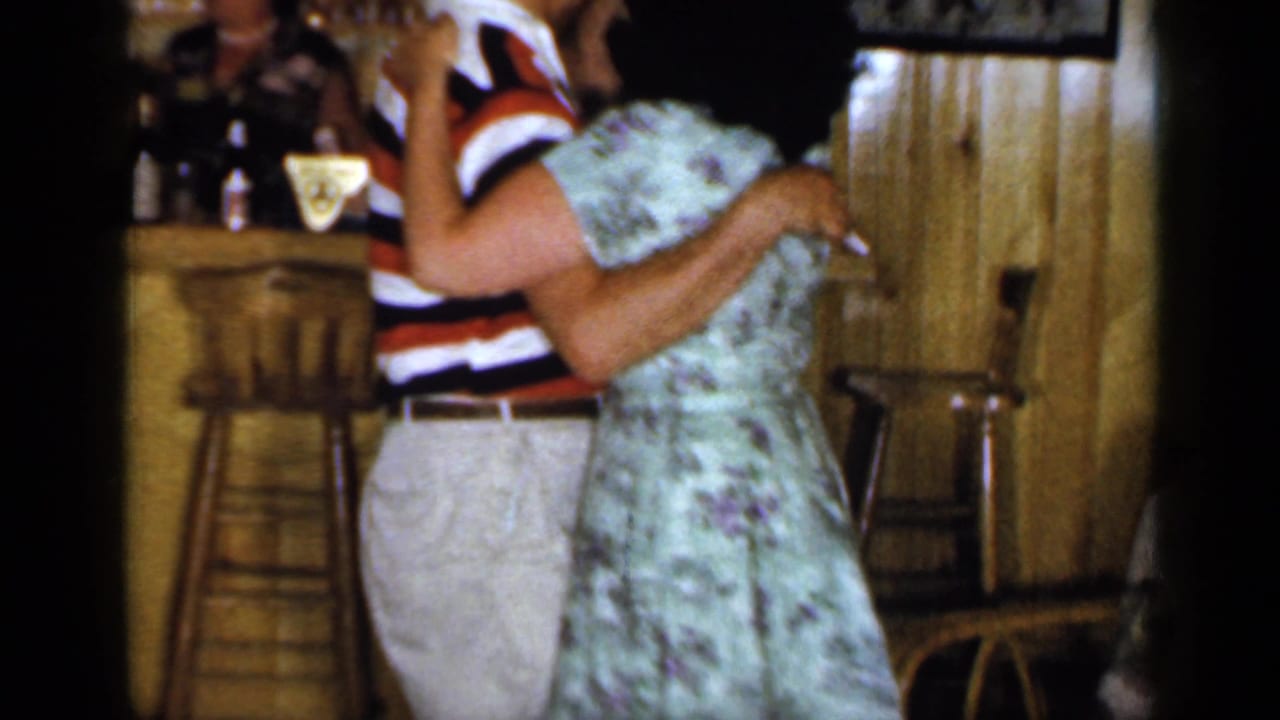 Man in striped shirt and woman in floral dress dance closely beside a bar.