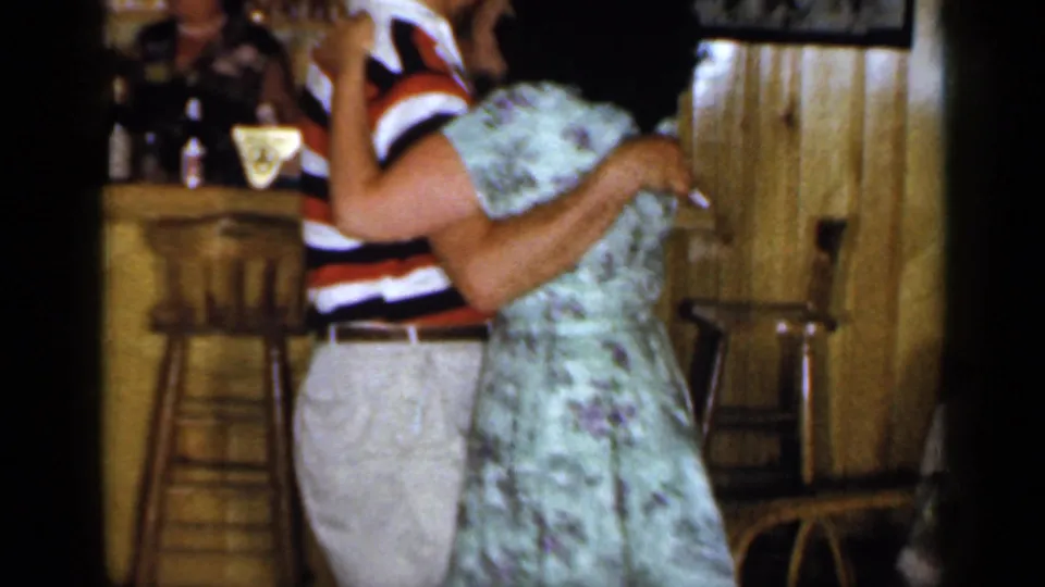 The couple sways beside the wood bar, the man in a red, white, and dark striped shirt and the woman in a light floral dress.