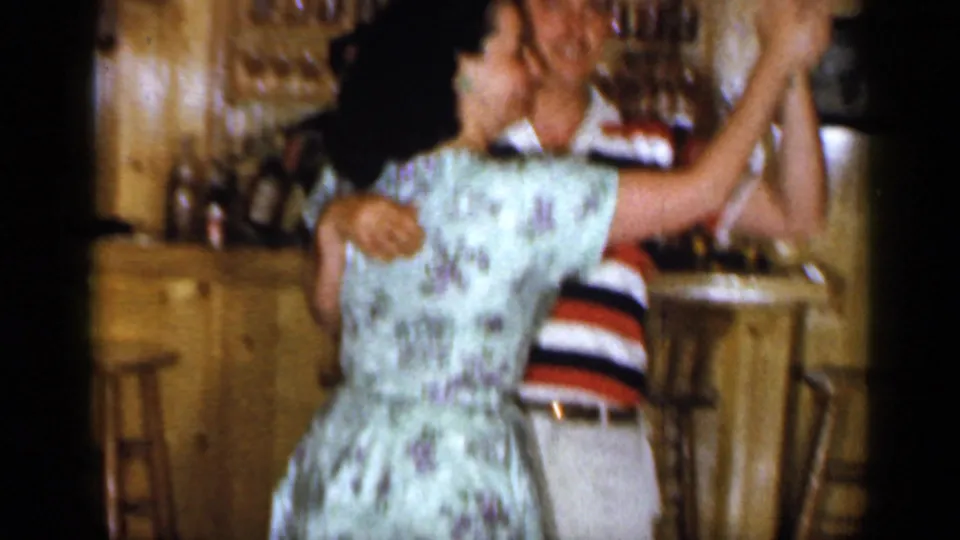 The woman in a pale floral dress faces away from the camera while dancing with the striped-shirted man in the warm bar interior.