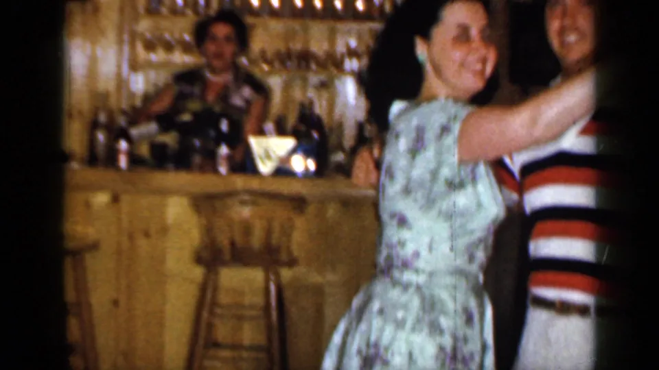 A smiling woman in a floral dress turns toward the camera as she dances, with a person visible behind the wood bar in the background.