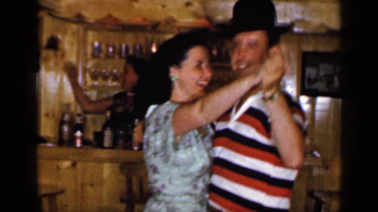 Smiling couple dancing in a wood-paneled basement bar with bottles behind them.