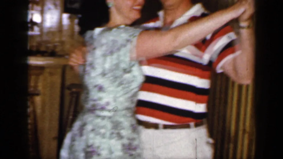 The couple continues dancing in the 1965 Long Island basement bar, with the man's black top hat and striped shirt prominent.