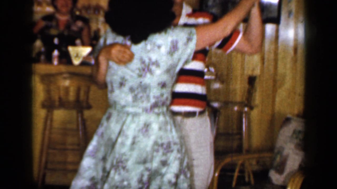 Woman in printed dress turns while dancing with a man in a striped shirt and dark hat.