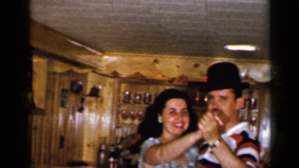 A smiling woman in a light blue printed dress faces the camera while dancing with a man in a bowler hat in 1965 Long Island footage.