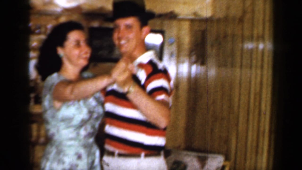 Man in striped shirt and dark hat dances with woman in floral dress inside a wood-paneled bar.