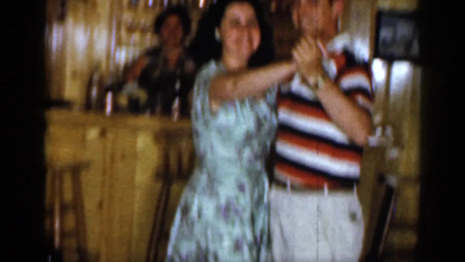 A woman in a light floral dress dances close to a man in a striped shirt while another adult is visible near the bar in the background.