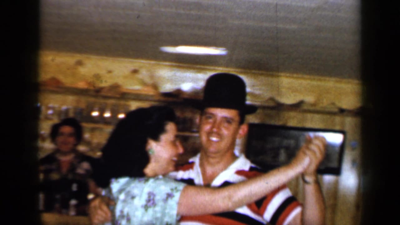 Man in a black hat dances with a woman in a floral dress inside a wood-paneled room.