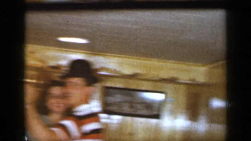 A blurred 1965 Long Island home-movie frame shows the man in a hat and striped shirt dancing with the woman near wood-paneled walls.