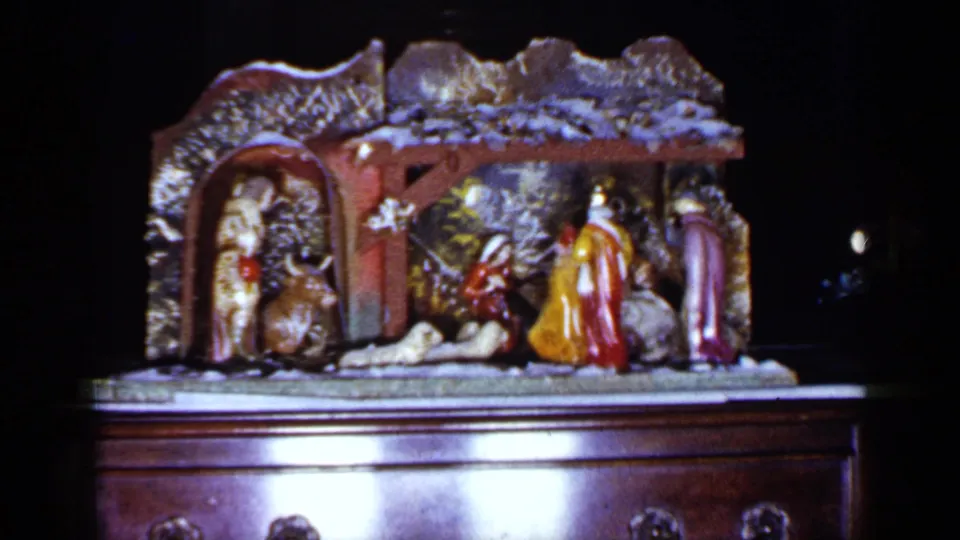 1965 Long Island home-movie frame showing a ceramic nativity manger display on a polished wooden cabinet.
