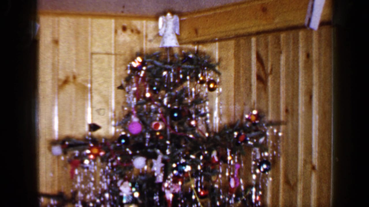 Tall Christmas tree with ornaments, tinsel, and angel topper near paneled wall.