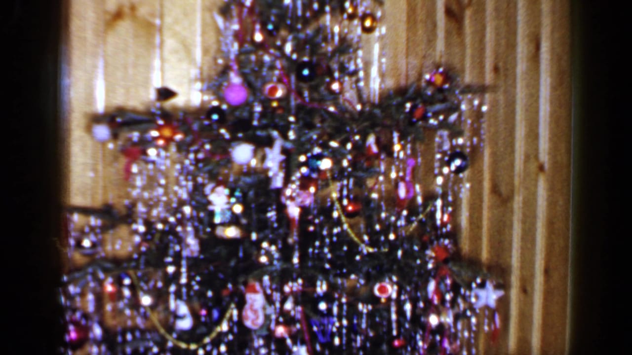 Christmas tree with bright ornaments, tinsel, and lights against wood paneling.