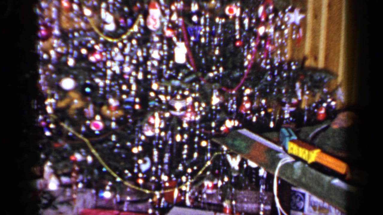 1965 Long Island home-movie frame of a glittering Christmas tree with ornaments, lights, and a long wrapped item beside it.