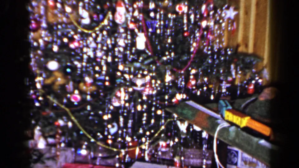 1965 Long Island home-movie frame of a glittering Christmas tree with ornaments, lights, and a long wrapped item beside it.