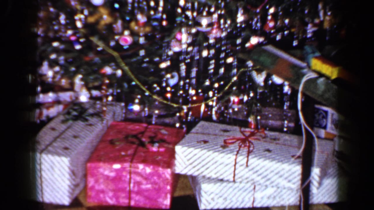 1965 Long Island home-movie frame of Christmas gifts under a tinsel tree, with white wrapped boxes, red bows, and a package at right.