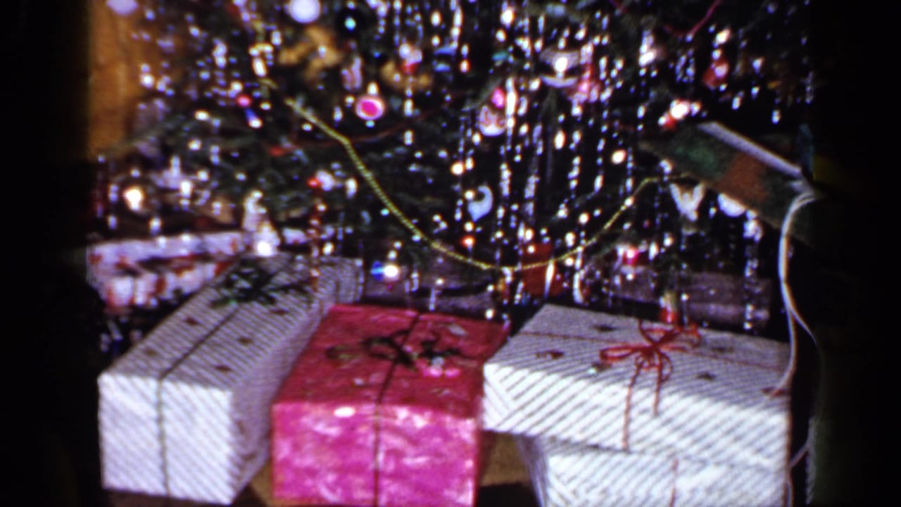 1965 Long Island home-movie frame showing wrapped gifts beneath a tinsel-covered Christmas tree in a private interior.