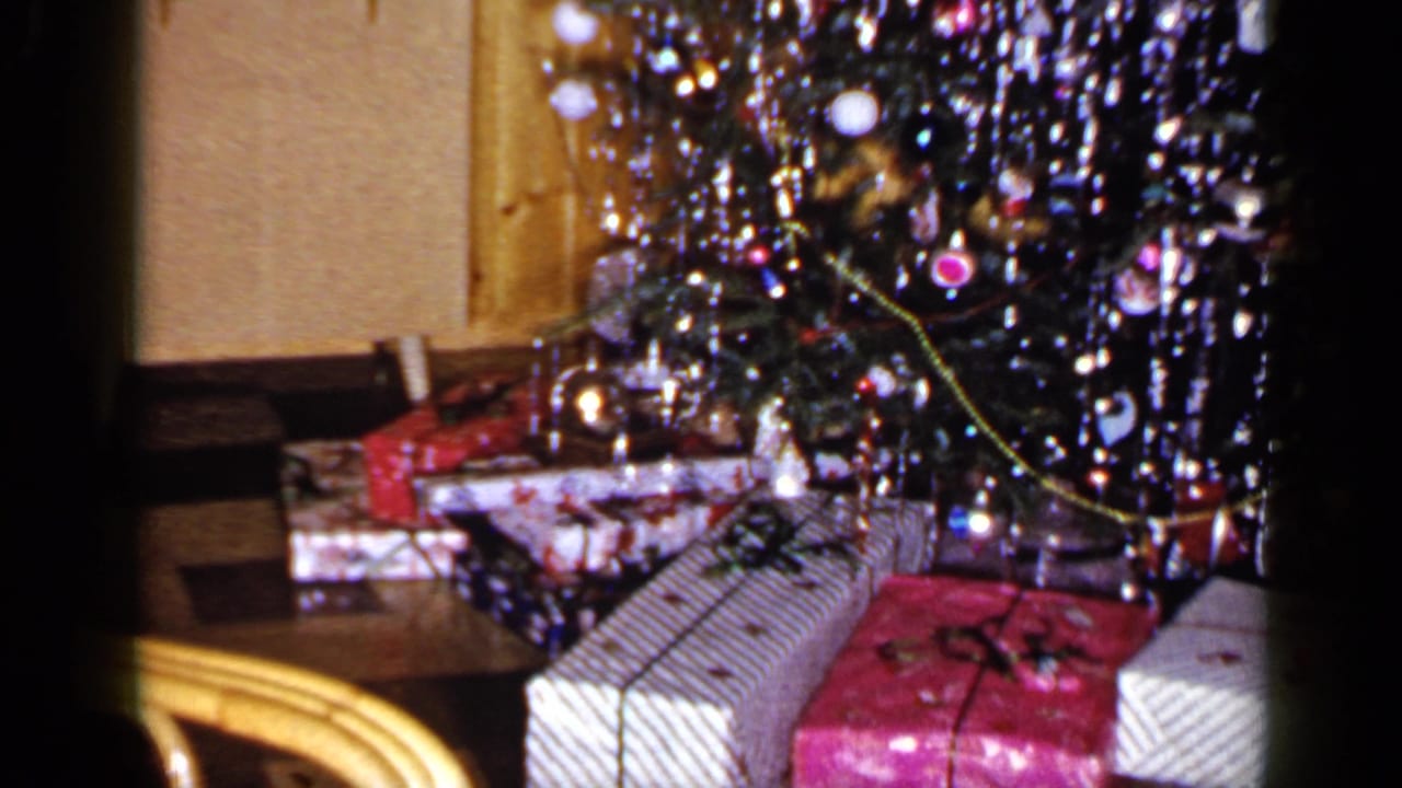 Home-movie view of a 1965 Long Island Christmas tree base with wrapped gifts, tinsel, ornaments, and a chair edge.