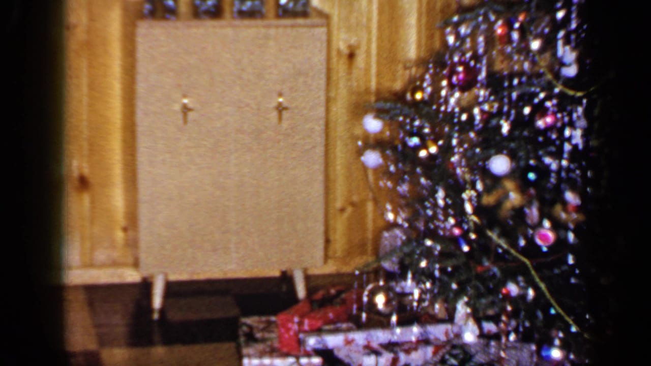 1965 Long Island home-movie frame showing a decorated Christmas tree, wrapped gifts and a small cabinet against wood paneling.