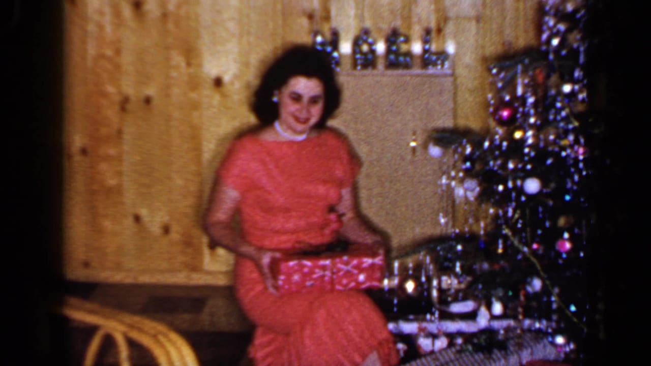 A woman in a red dress sits in a Long Island living room beside a tinsel-trimmed Christmas tree, holding a wrapped present.