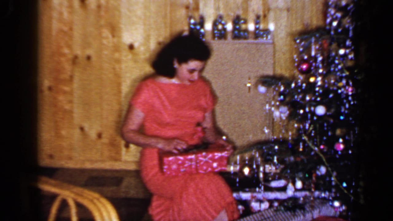 In the 1965 home-movie scene, the woman looks down and works at the wrapping paper near the decorated Christmas tree.