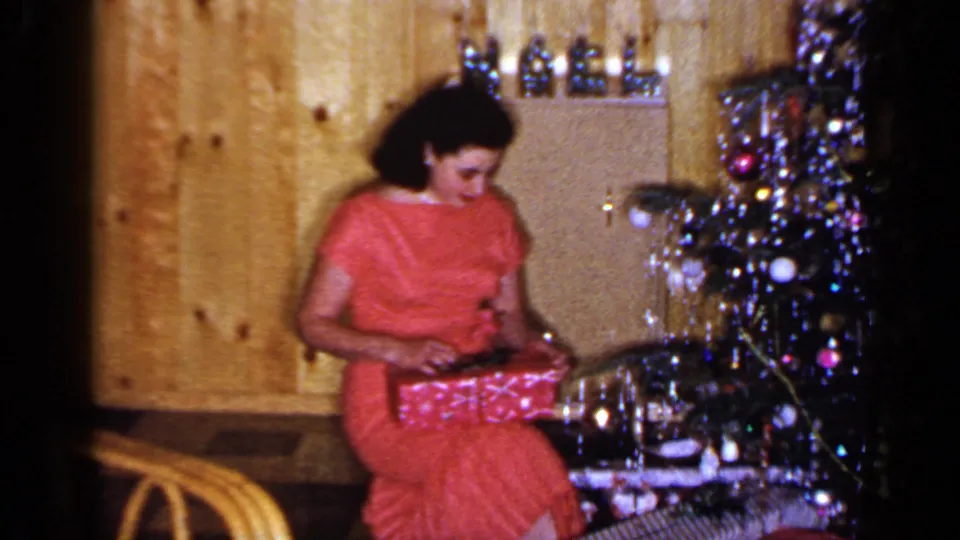 In the 1965 home-movie scene, the woman looks down and works at the wrapping paper near the decorated Christmas tree.