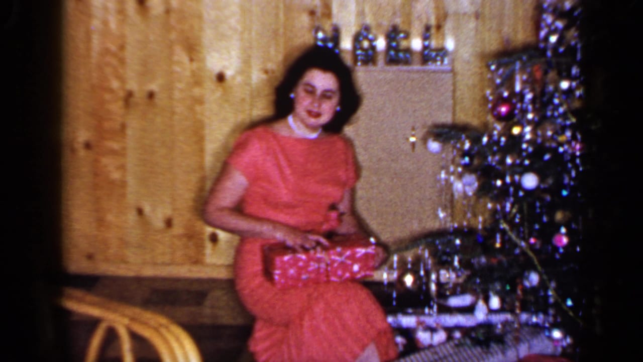 The woman in a red dress smiles toward the camera with the Christmas present on her lap in a wood-paneled Long Island interior.