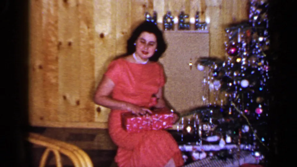 The woman in a red dress smiles toward the camera with the Christmas present on her lap in a wood-paneled Long Island interior.
