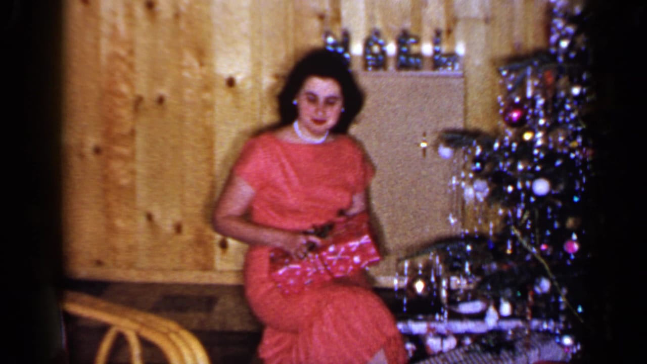 Woman in a red dress seated beside a Christmas tree in a wood-paneled room.