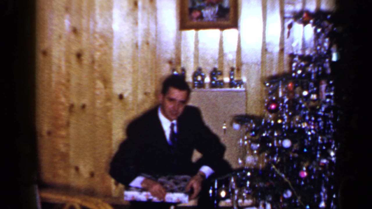 A man in a dark suit sits beside a decorated Christmas tree in a 1965 Long Island home interior, holding a present.