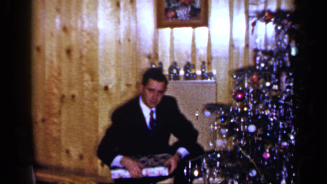 A 1965 Long Island frame shows a man in formal clothing seated with a wrapped gift near a decorated Christmas tree.