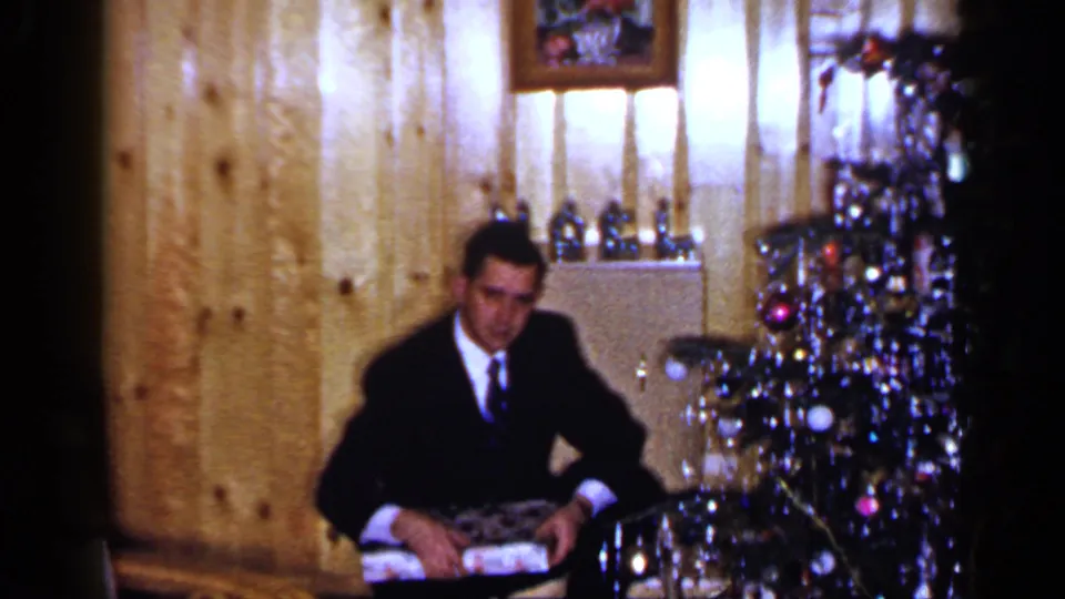 A 1965 Long Island frame shows a man in formal clothing seated with a wrapped gift near a decorated Christmas tree.