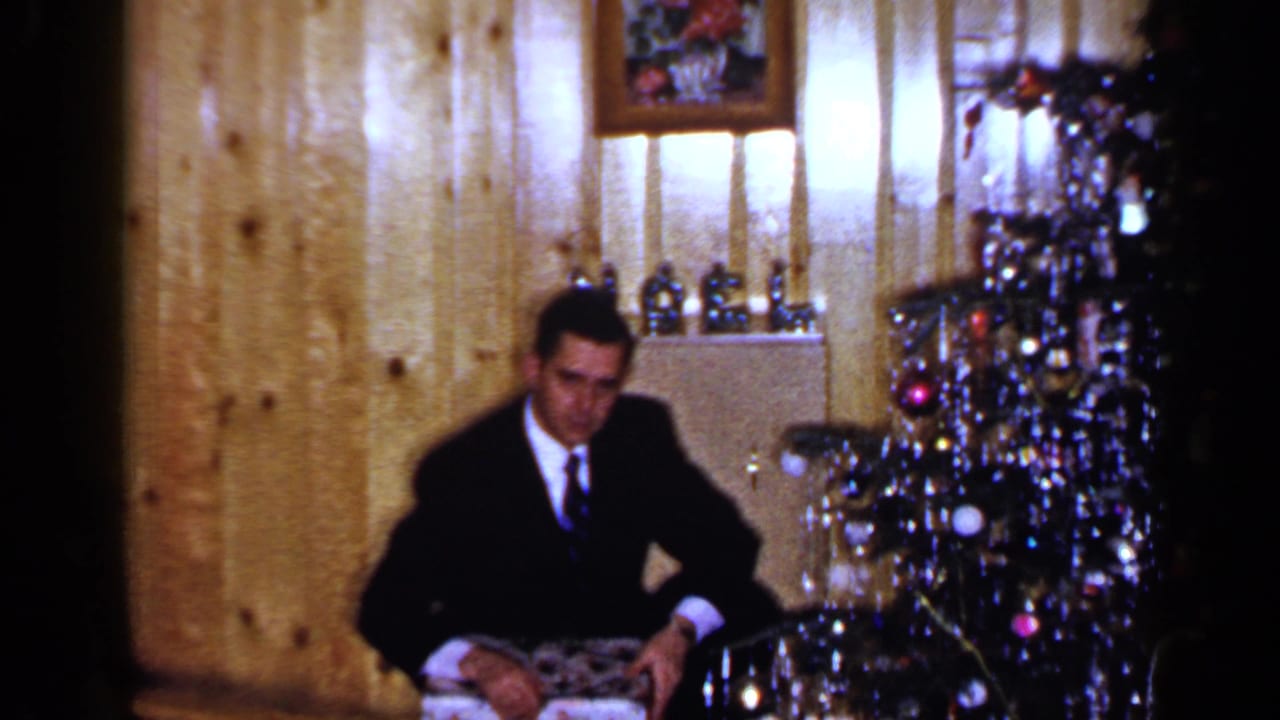 A man in a dark suit sits by the decorated Christmas tree with a wrapped gift in a 1965 Long Island home-movie frame.