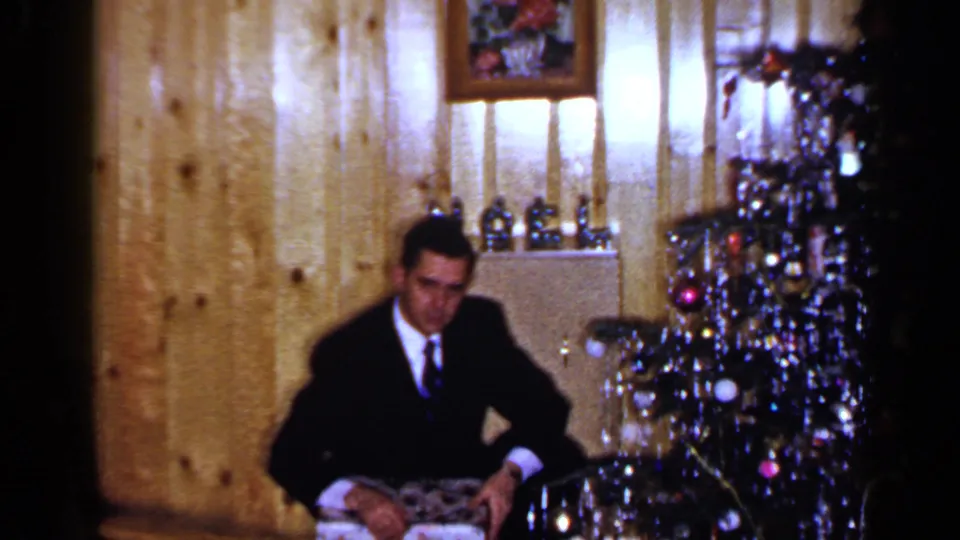 A man in a dark suit sits by the decorated Christmas tree with a wrapped gift in a 1965 Long Island home-movie frame.