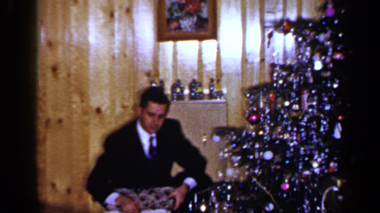 Man in a dark suit sits beside a decorated Christmas tree holding a wrapped gift.