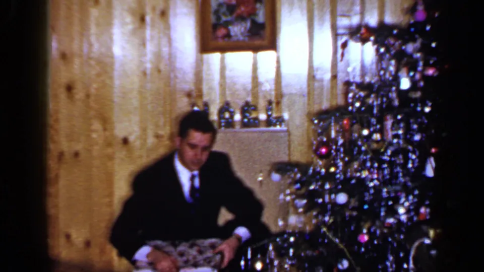 In a 1965 Long Island home-movie frame, a suited adult man sits near a tinsel Christmas tree and handles a wrapped gift.