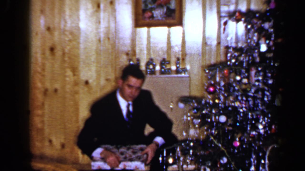Man in a dark suit sits on the floor near a Christmas tree with a gift in his lap.
