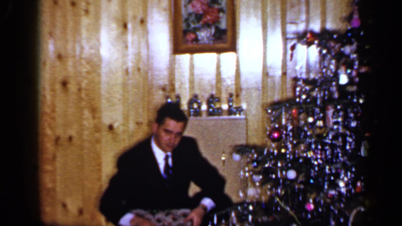 A soft 8mm frame shows a suited man in a private Long Island interior holding a patterned Christmas gift beside the tree.