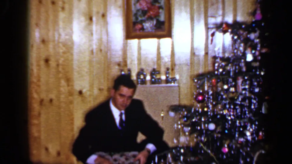 A soft 8mm frame shows a suited man in a private Long Island interior holding a patterned Christmas gift beside the tree.