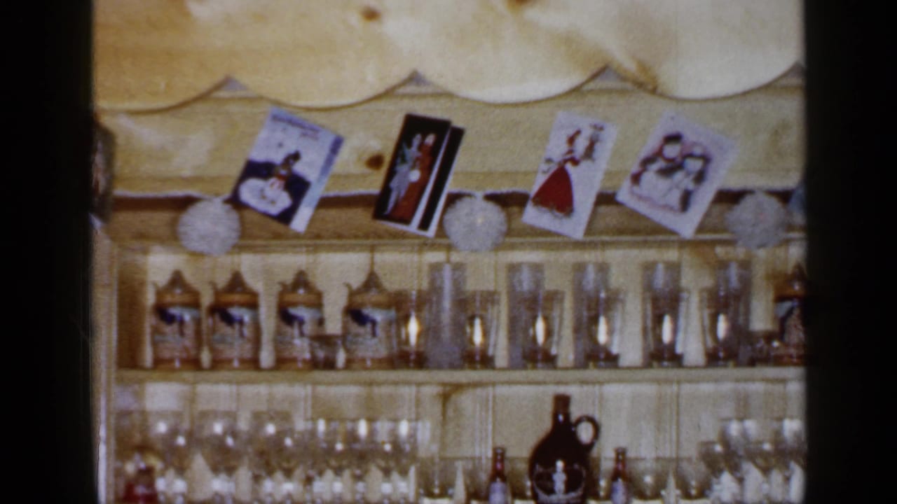 Long Island 1965 home-movie frame of a pine shelf loaded with steins, glassware, Christmas cards, and dark ceramic jugs.