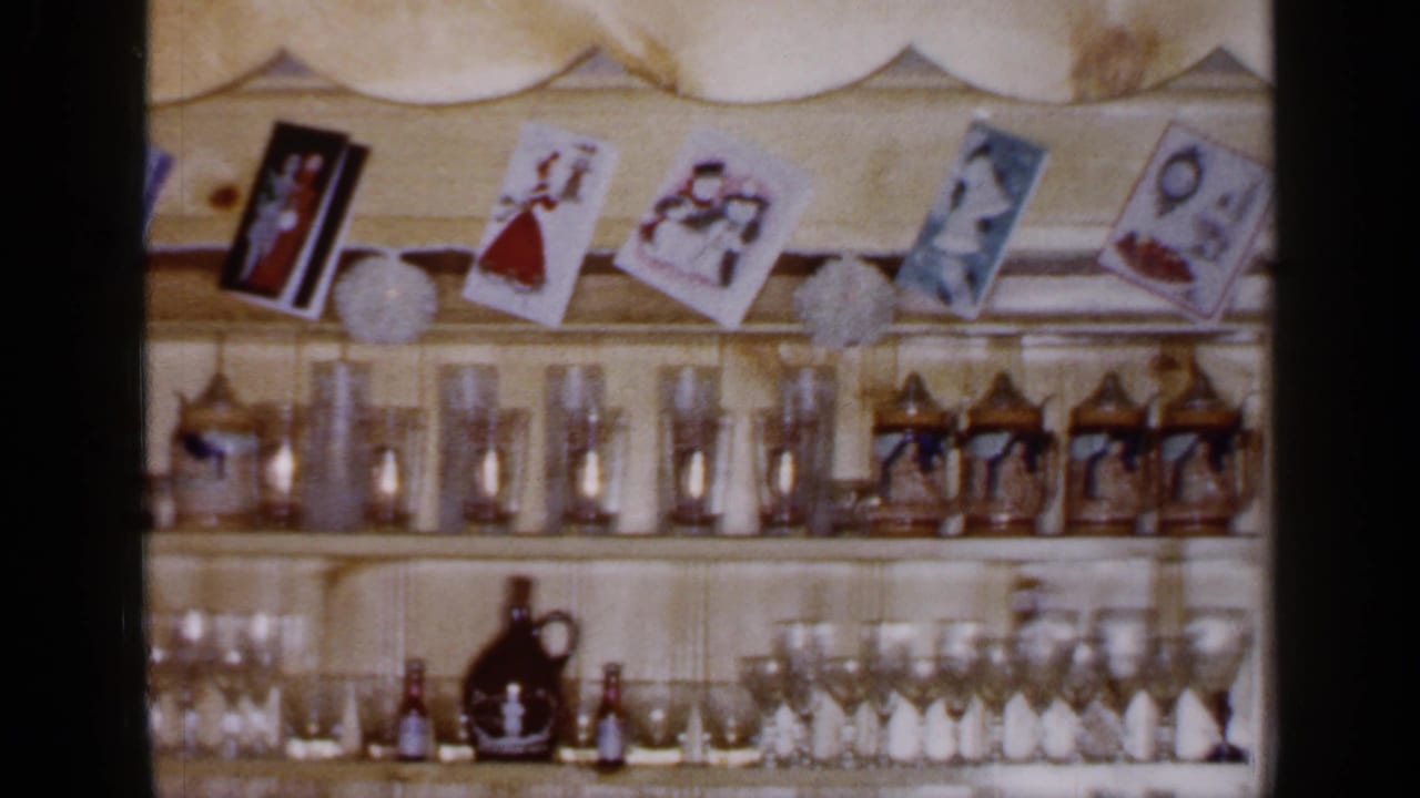 1965 Long Island home-movie frame showing a Christmas wall shelf with cards, white garland, glassware, steins, and small bottles.