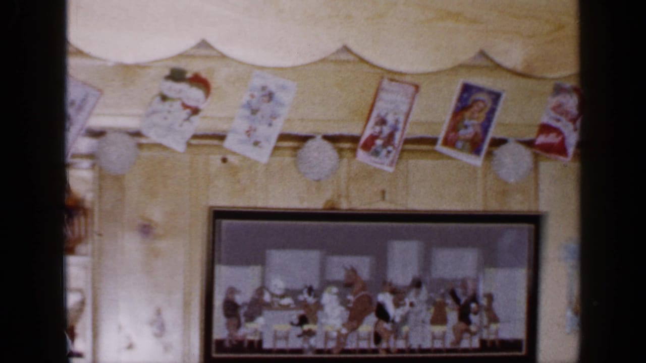 1965 Long Island home-movie frame showing Christmas cards and white ornaments strung above a framed holiday picture on a paneled wall.