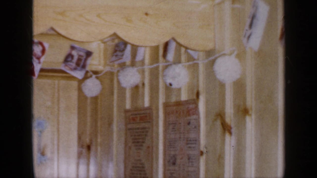 Final 1965 Long Island home-movie frame showing the right side of a Christmas card garland and posted papers on a paneled wall.