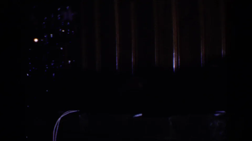 Dim 1965 Long Island home-movie frame showing a faint decorated Christmas tree beside vertical wood paneling.