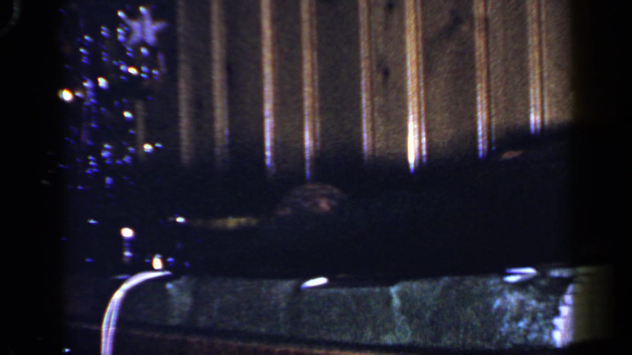 1965 Long Island holiday home-movie frame with a decorated tree, dim furniture, and wood-paneled interior background.