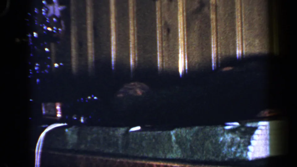 1965 Long Island home-movie frame showing a toy train setup indoors, with wood paneling, shadowed track scenery, and a Christmas tree at left.