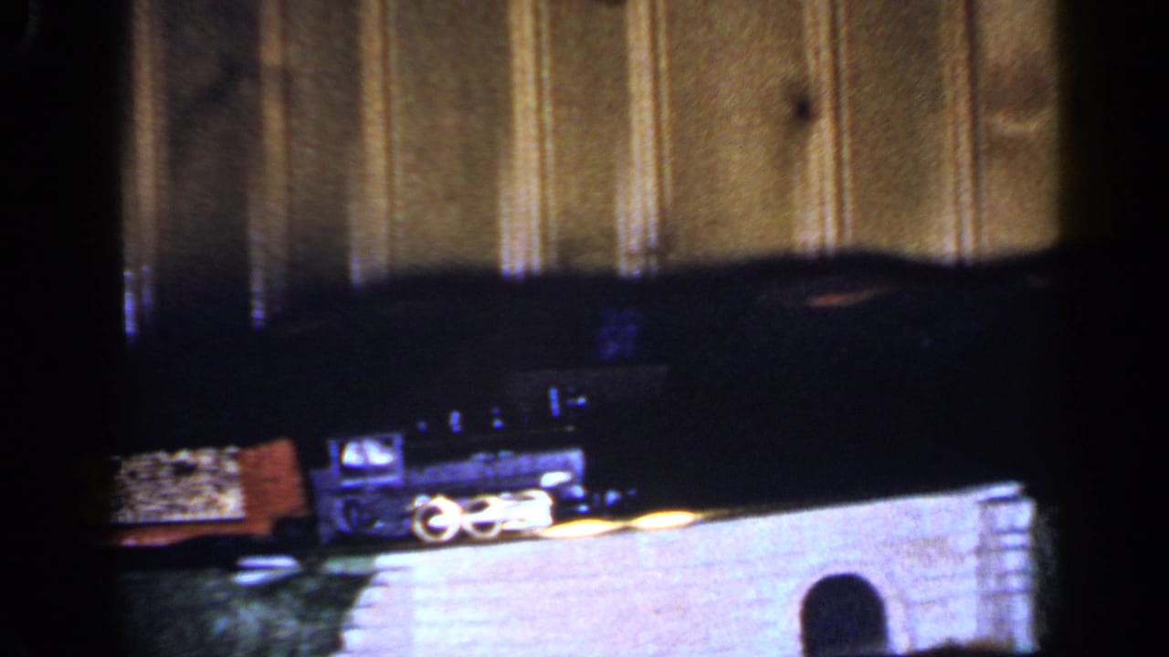 A 1965 Long Island home-movie frame shows small toy train cars and track scenery indoors, with wood paneling and a white wall element behind them.