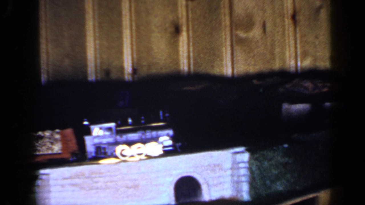 1965 Long Island 8mm frame showing a black toy train and cars moving across a small bridge against wood paneling.