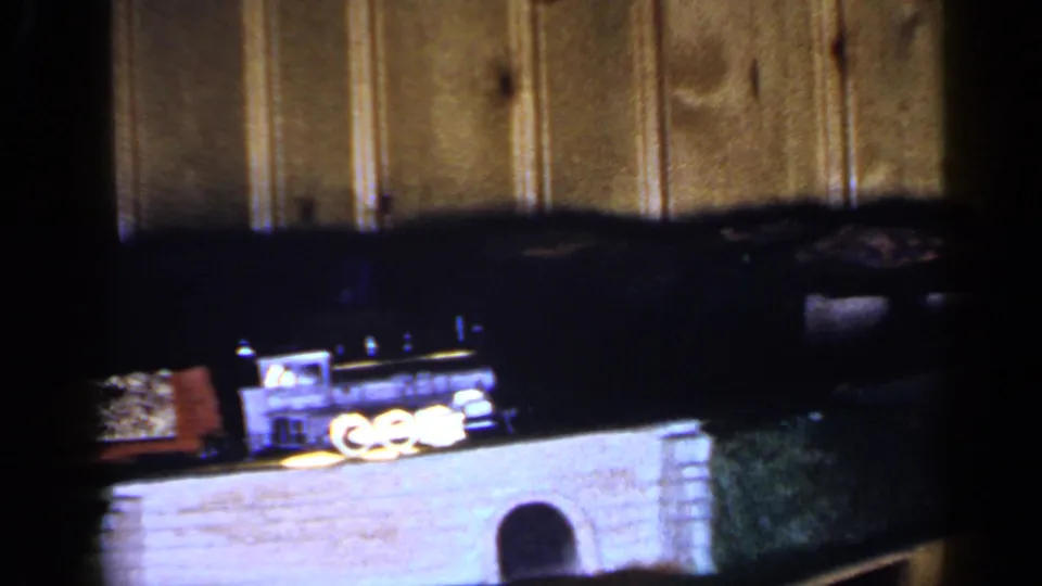 1965 Long Island 8mm frame showing a black toy train and cars moving across a small bridge against wood paneling.